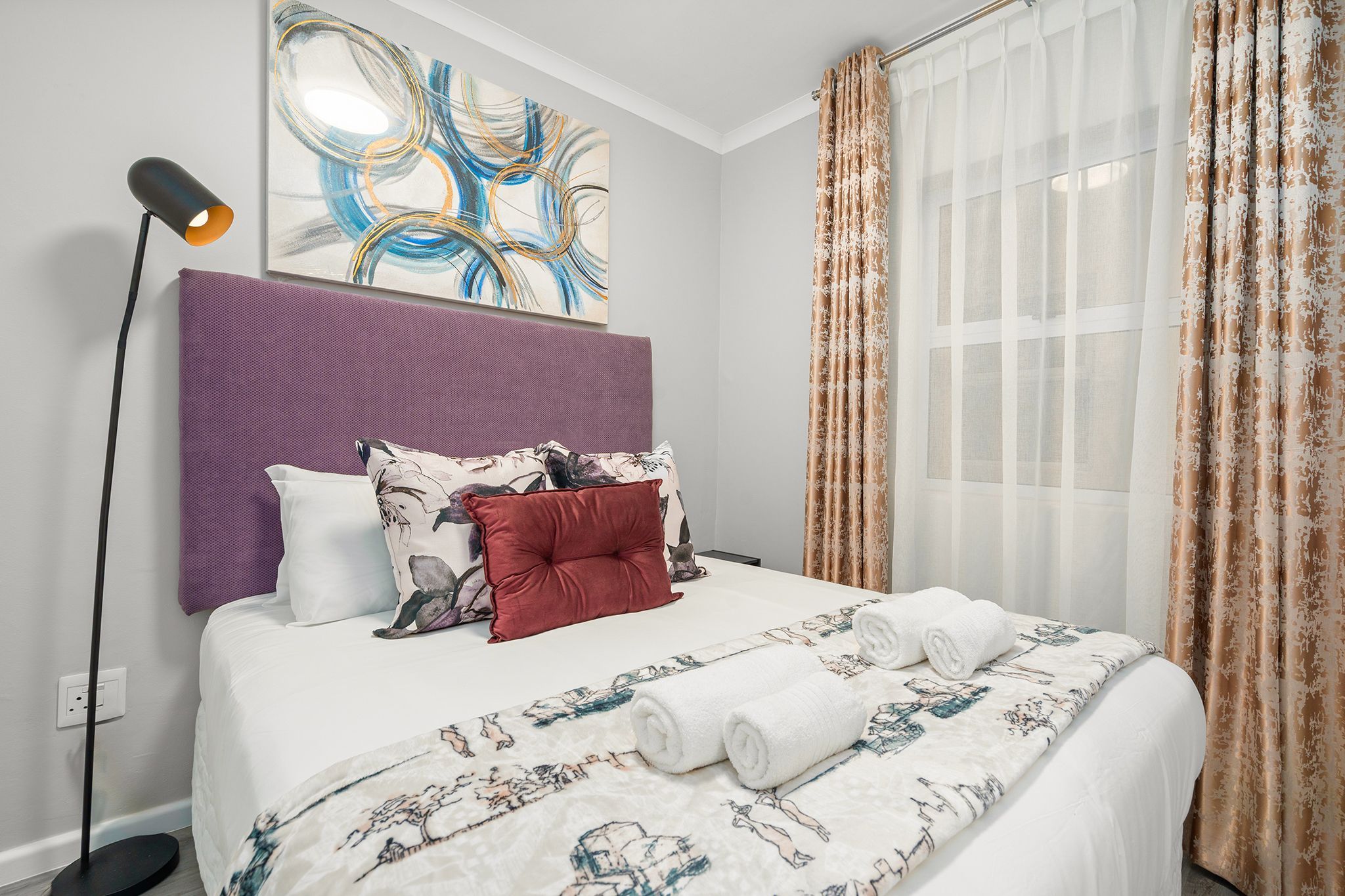 A neatly made bed with a floral bedspread and two rolled towels, a purple headboard, and a modern abstract painting above the bed.
