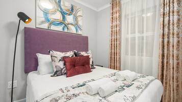 A neatly made bed with a floral bedspread and two rolled towels, a purple headboard, and a modern abstract painting above the bed.