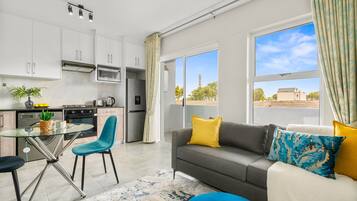 A modern kitchen with white cabinets, a glass dining table, and a sofa with yellow and blue cushions.