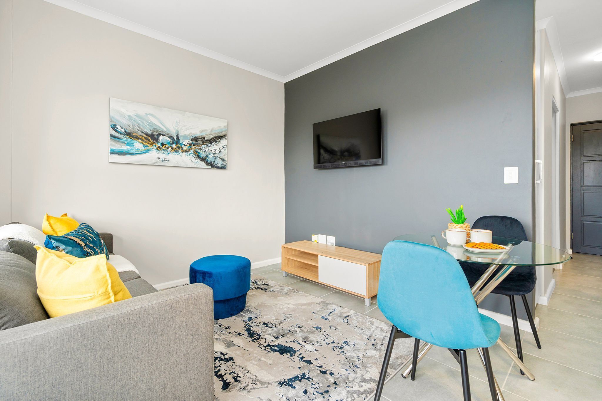A modern living room with a grey sofa, blue and yellow cushions, a glass coffee table, and a blue chair.