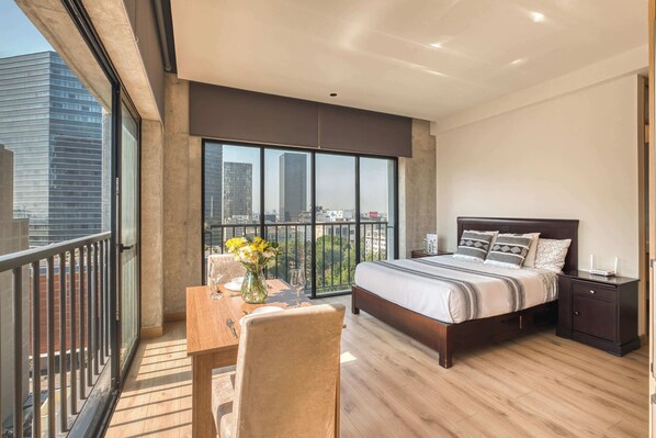 Une chambre moderne avec un grand lit, une table de chevet, une commode, une table à manger avec des chaises, et une vue sur la ville grâce à de grandes fenêtres.