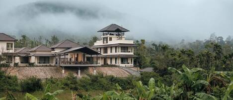 A multi-story house with a balcony, surrounded by lush greenery and a misty mountain backdrop.