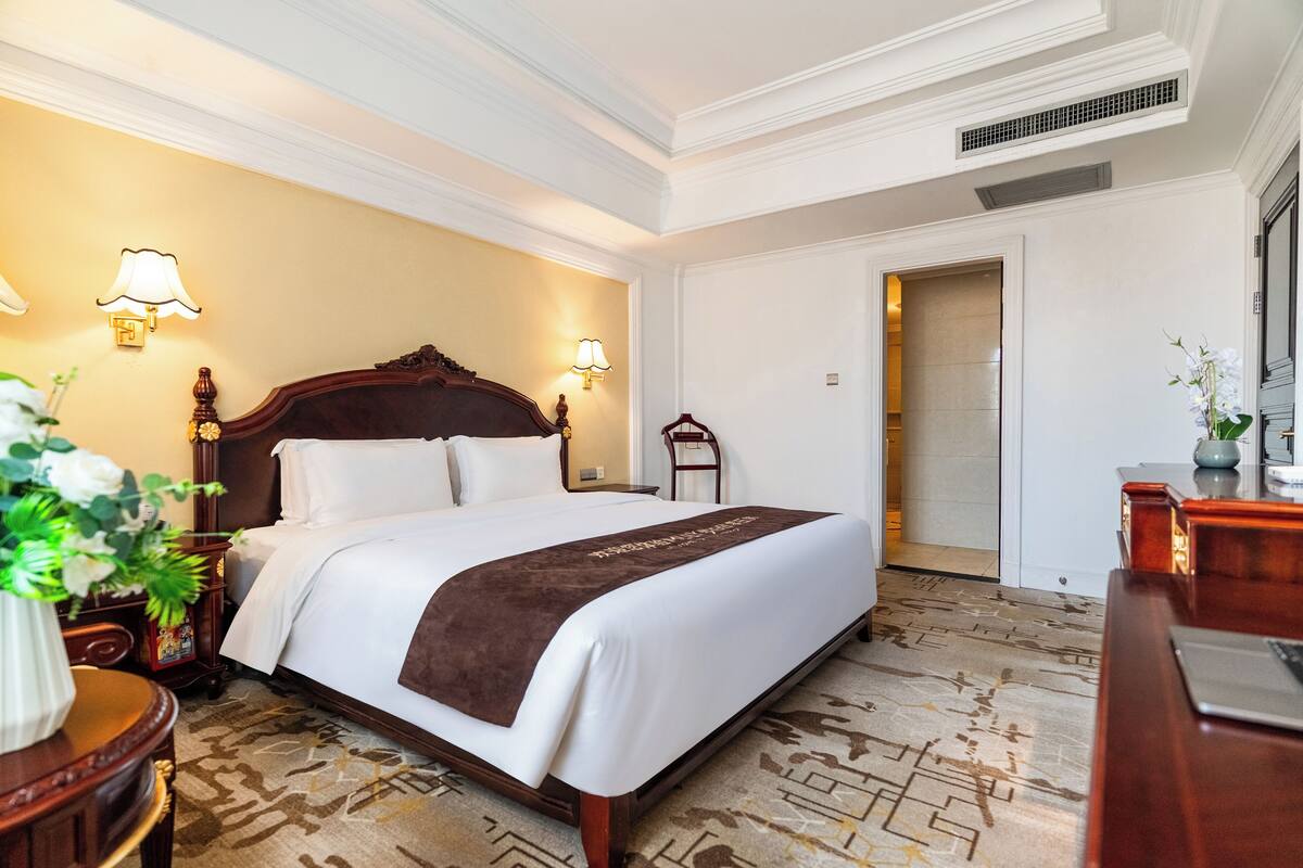 Habitación de hotel con una cama grande, dos lámparas de noche, un cabecero de madera, una mesita de noche con un jarrón de flores y una puerta que da a otra habitación.