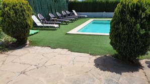 Une terrasse en pierre menant à un espace piscine agrémenté de gazon synthétique et de chaises longues.