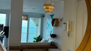 Un interior moderno con un espejo redondo que refleja una habitación con una araña, una planta en maceta y vistas al mar a través de una ventana.