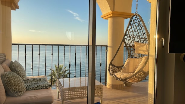 Un balcón con una silla colgante, un sofá con cojines estampados y vistas al mar.