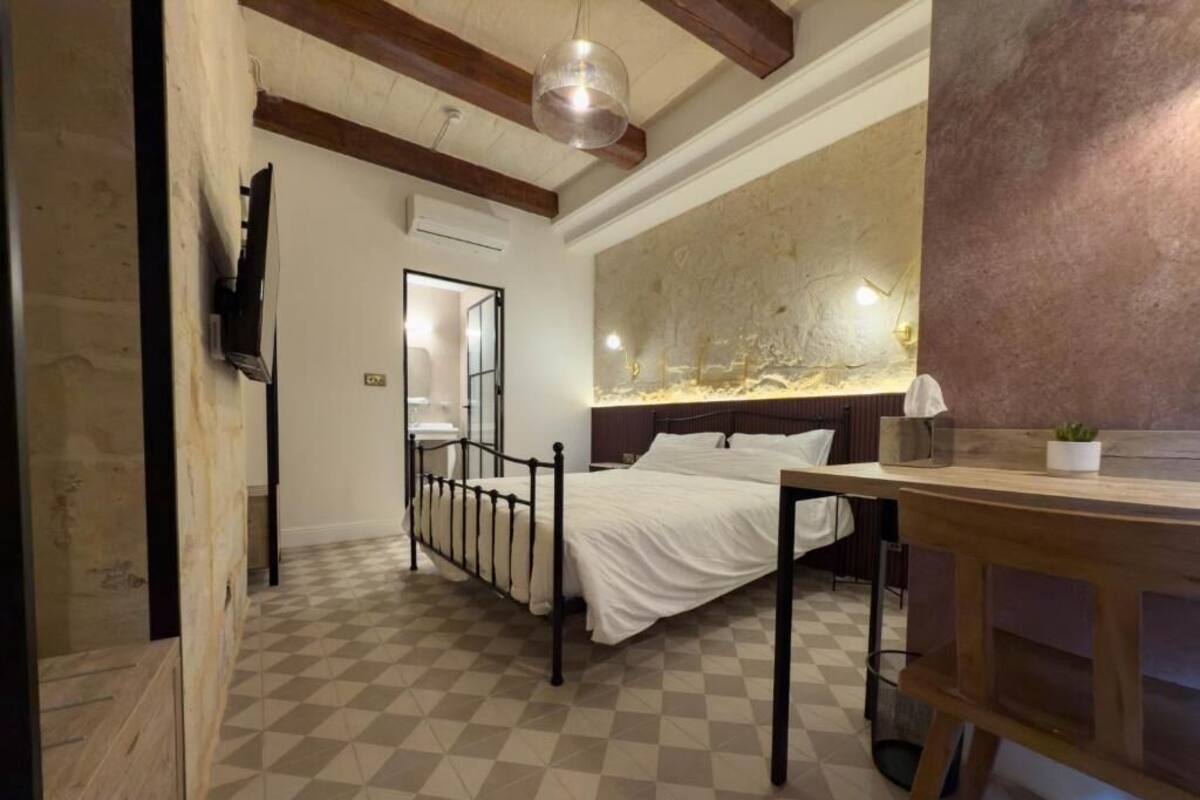 A bedroom with a checkered floor, a bed with white bedding, a wooden table, and a large abstract wall art piece.