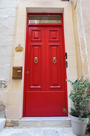 A red double door with a brass knocker, a mailbox, and a potted plant beside it. - Fourteen Guesthouse (Żejtun)