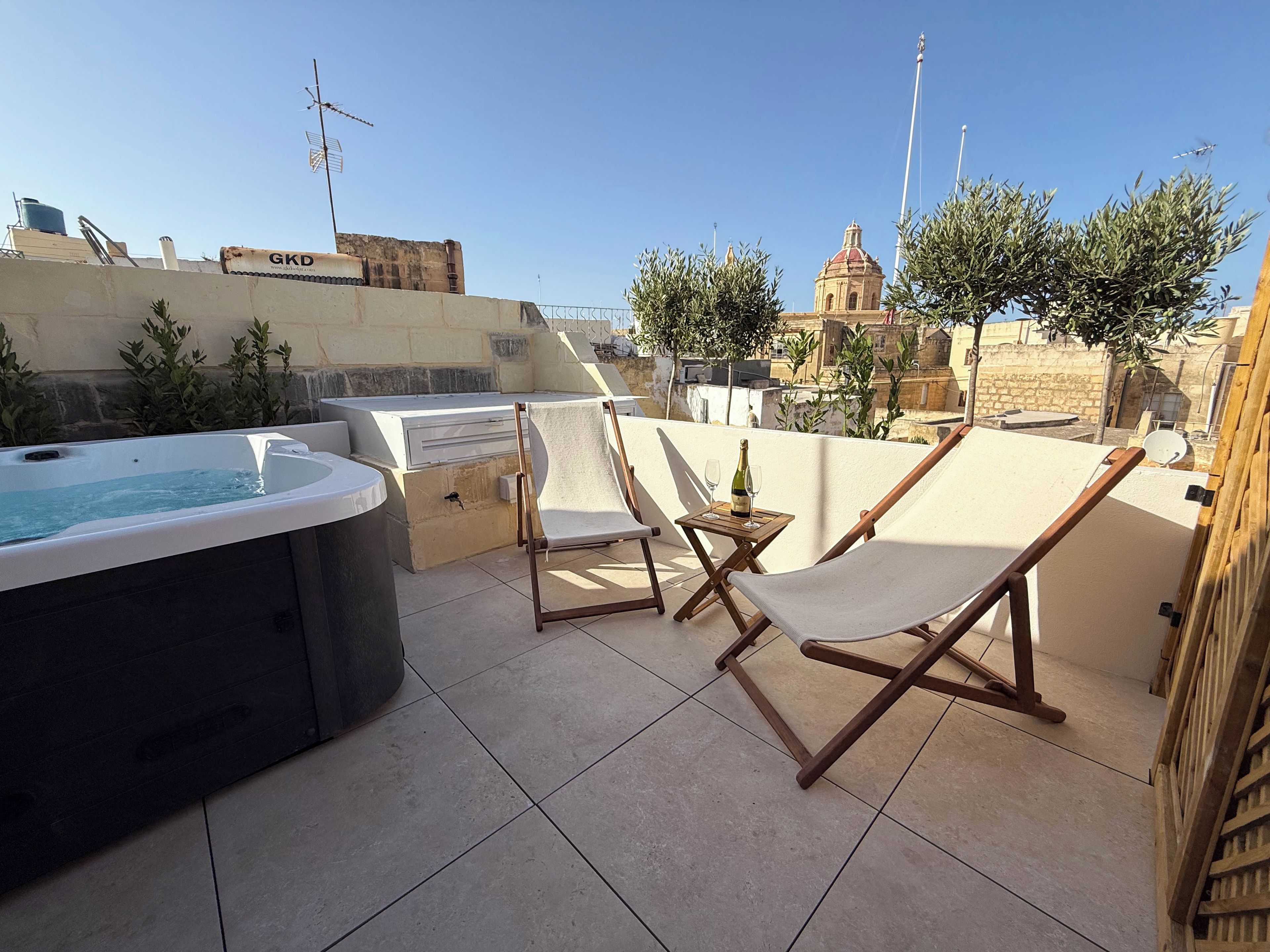 A rooftop terrace with a hot tub, two lounge chairs, and a small table with a bottle and glasses.