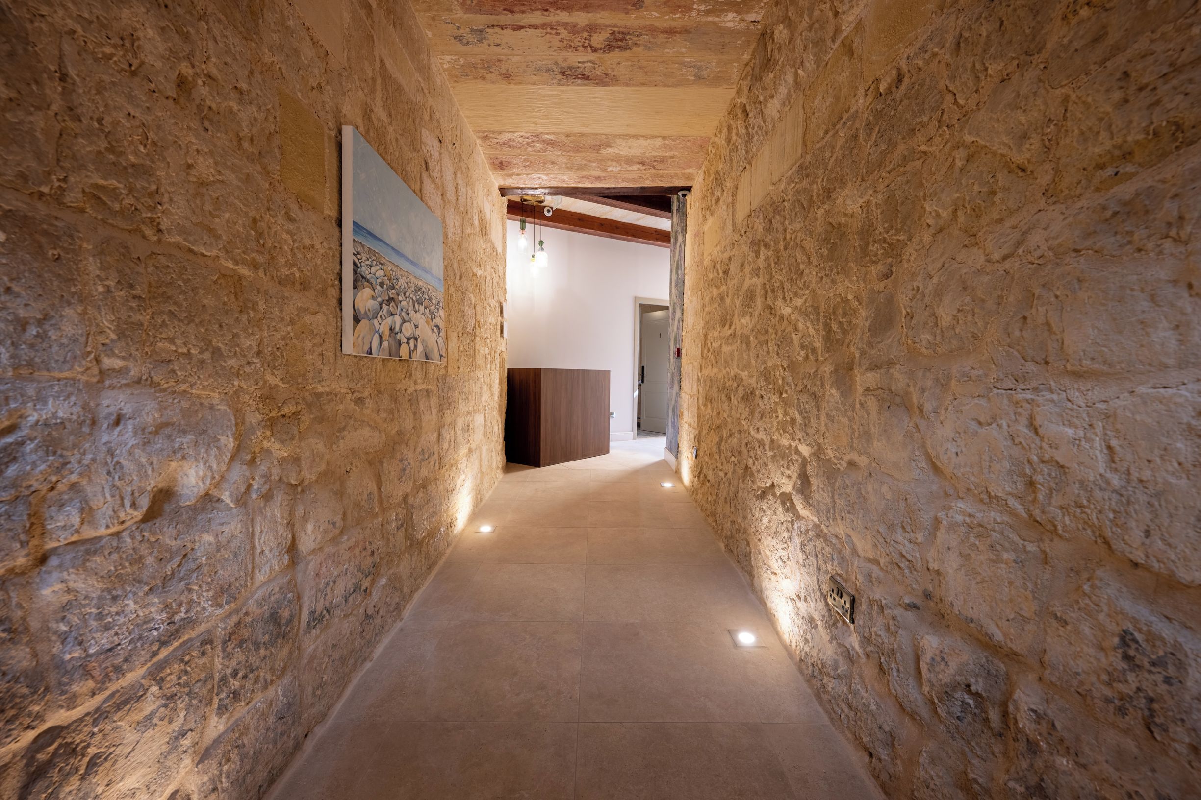 A narrow stone corridor with a painting on the left wall, leading to a room with a wooden door.