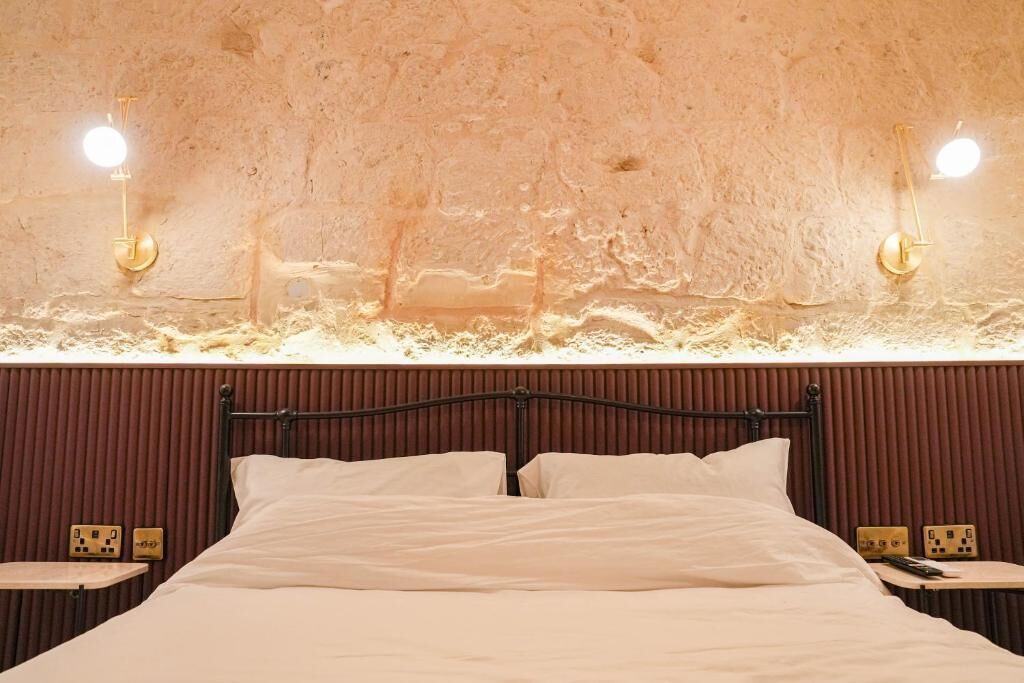 Una cama con cabecera, dos mesitas de noche e iluminación de pared.