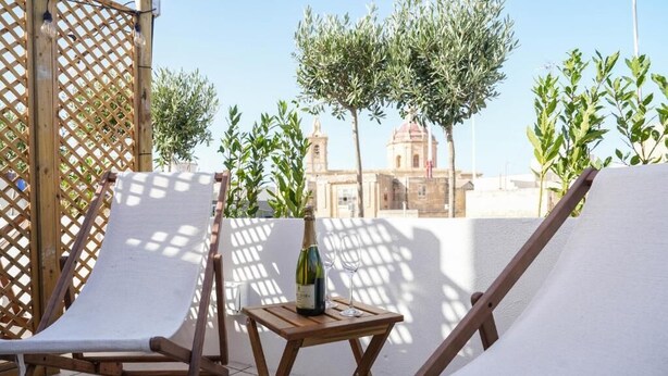 A terrace with two lounge chairs, a small wooden table with a bottle and glass, and a view of a building with a dome in the background. - Fourteen Guesthouse  (Żejtun)