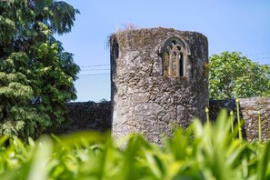 A stone tower with arched windows, surrounded by greenery and a fence.