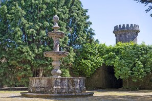 A stone fountain with a balustrade, surrounded by trees and a stone tower in the background.