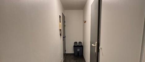 A narrow hallway with a carpeted floor, white walls, and a ceiling light. There are two doors on the right side and a small trash bin in the center.