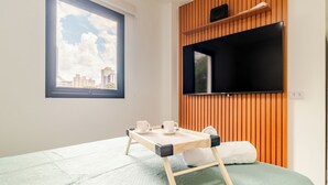 Ein Hotelzimmer mit Bett, Fernseher an einer Wand aus Holzpaneelen, einem Fenster mit Blick auf die Stadt und einem Tablett mit Tassen und einem Buch.