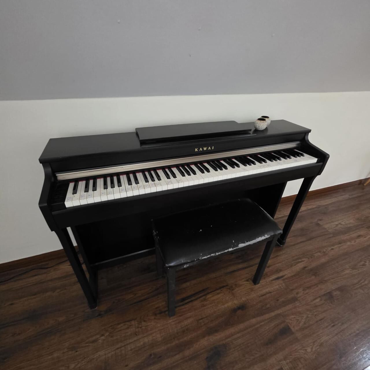 Un piano vertical negro con un banco, situado frente a una pared blanca sobre un piso de madera.