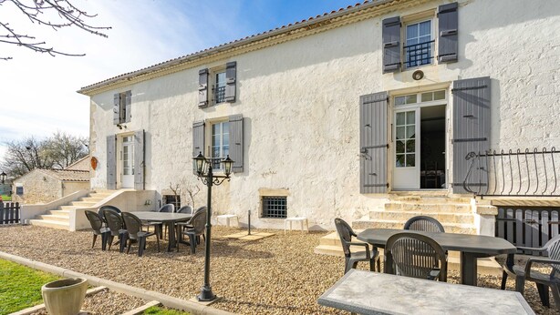 A white building with a stone facade, outdoor seating area with tables and chairs, and a gravel path.