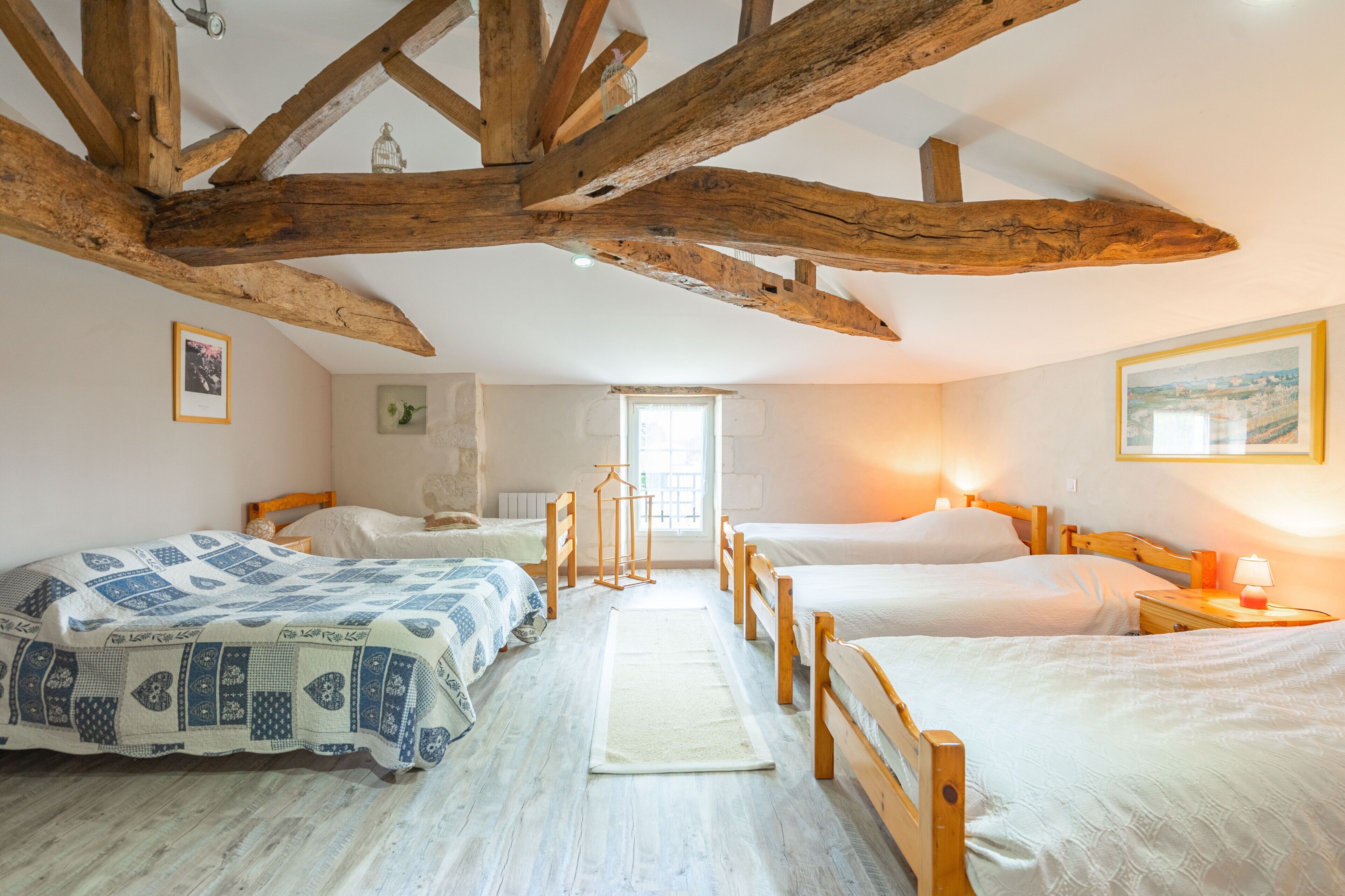 A bedroom with four beds arranged in a row, wooden beams on the ceiling, and a window with natural light.