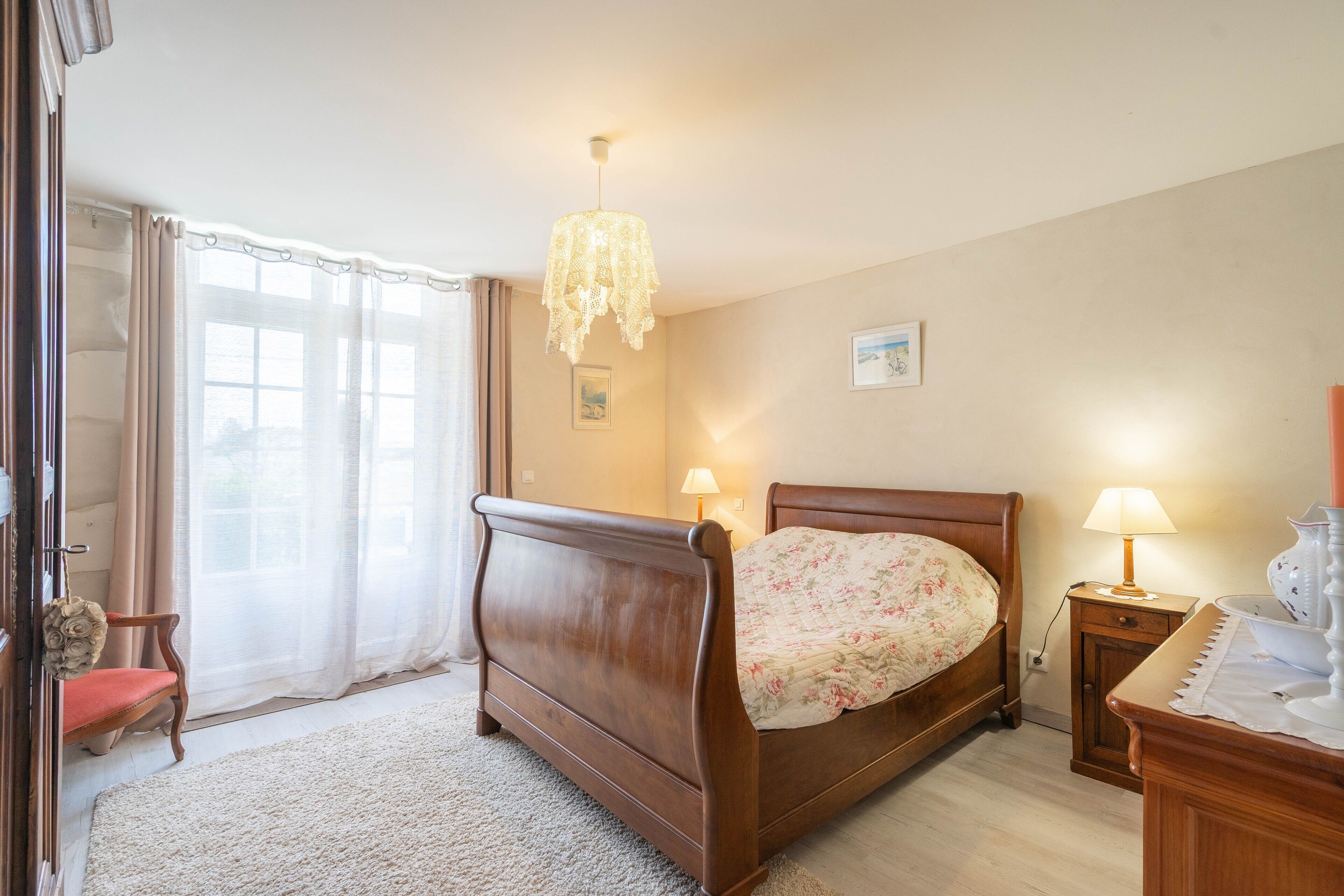 A bedroom with a wooden bed, a nightstand with a lamp, a chair, a window with curtains, and a chandelier.