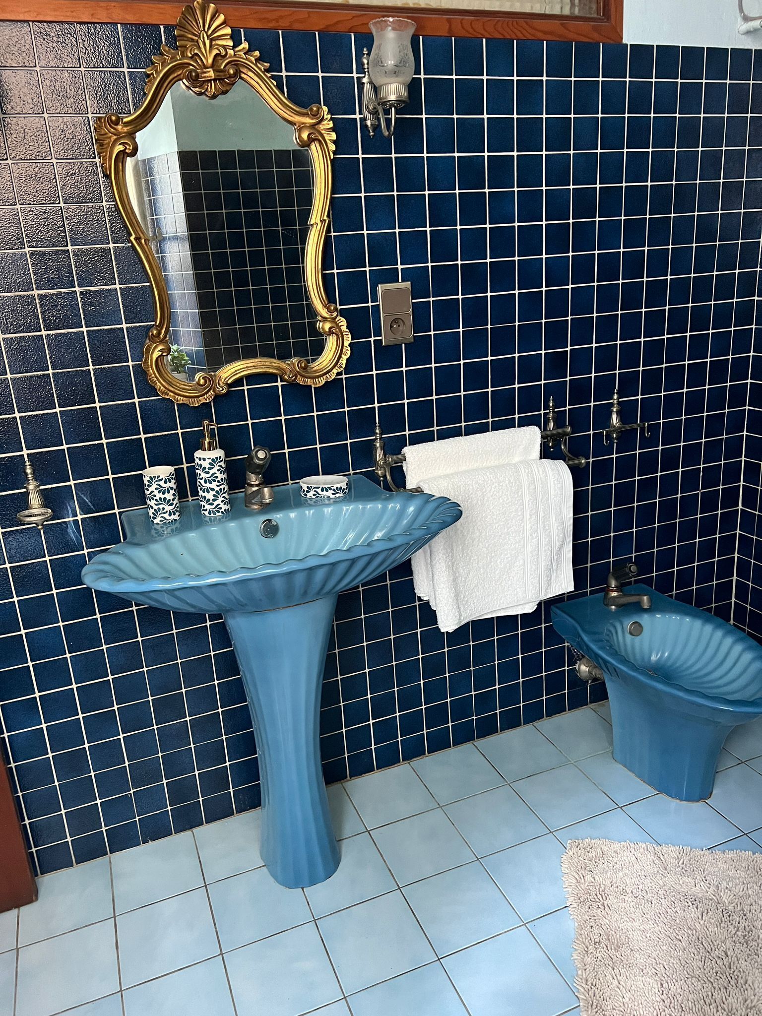 Une salle de bain avec un mur recouvert de carreaux bleus, un grand miroir avec un cadre doré, un lavabo sur colonne, un bidet et un porte-serviettes.