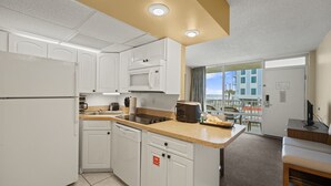A compact kitchen with white cabinets, a microwave, a toaster, and a coffee maker. There is a small dining area with a table and chairs, and a view of the beach through large windows. - Easy Oceanfront Escape | 1BR Sleeps 6 Ormond Beach (Ormond Beach)