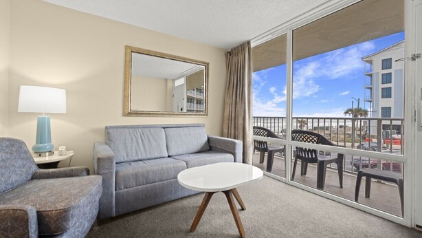 A hotel room with a grey sofa, a small round table, a lamp, and a large window with a balcony. - Easy Oceanfront Escape | 1BR Sleeps 6 Ormond Beach (Ormond Beach)