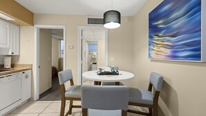 Cozinha moderna com armários brancos, sala de jantar com mesa redonda e duas cadeiras, e uma grande pintura abstrata na parede.