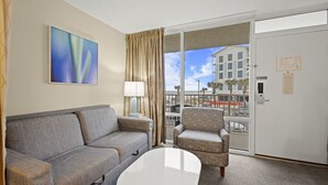 A hotel room with a grey sofa, a patterned armchair, a white coffee table, and a large window with a view of a building and palm trees.
