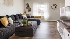 A modern living room with a sectional sofa, a coffee table with decorative items, a TV, and a dining area with a table and chairs. - Home-away-from-home, Entire home in West Des Moines, Iowa (West Des Moines)