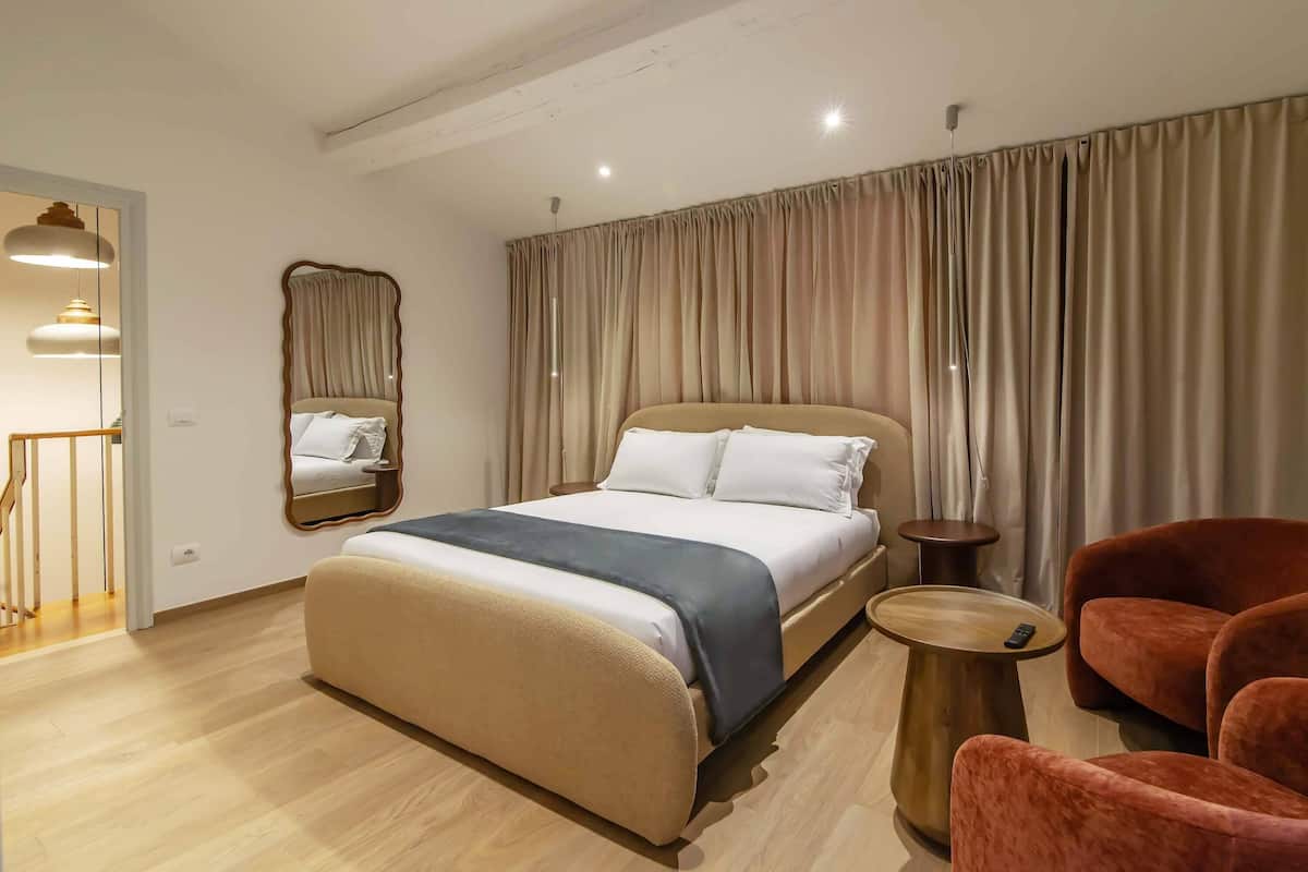 Una habitación de hotel moderna con una cama grande, un área de descanso con sofá y sillones, una mesita auxiliar y un espejo en la pared.