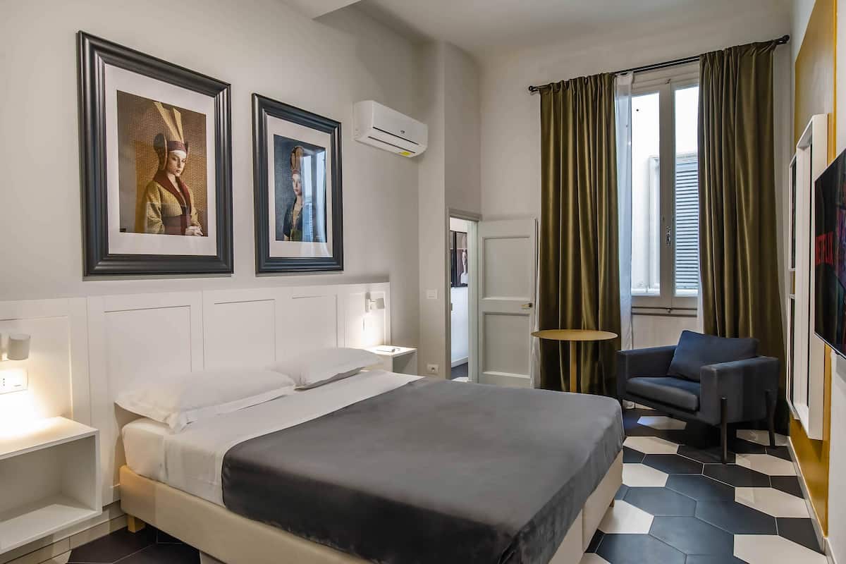 A hotel room with a bed, two framed pictures, an air conditioner, a chair, a TV, and a window with curtains.