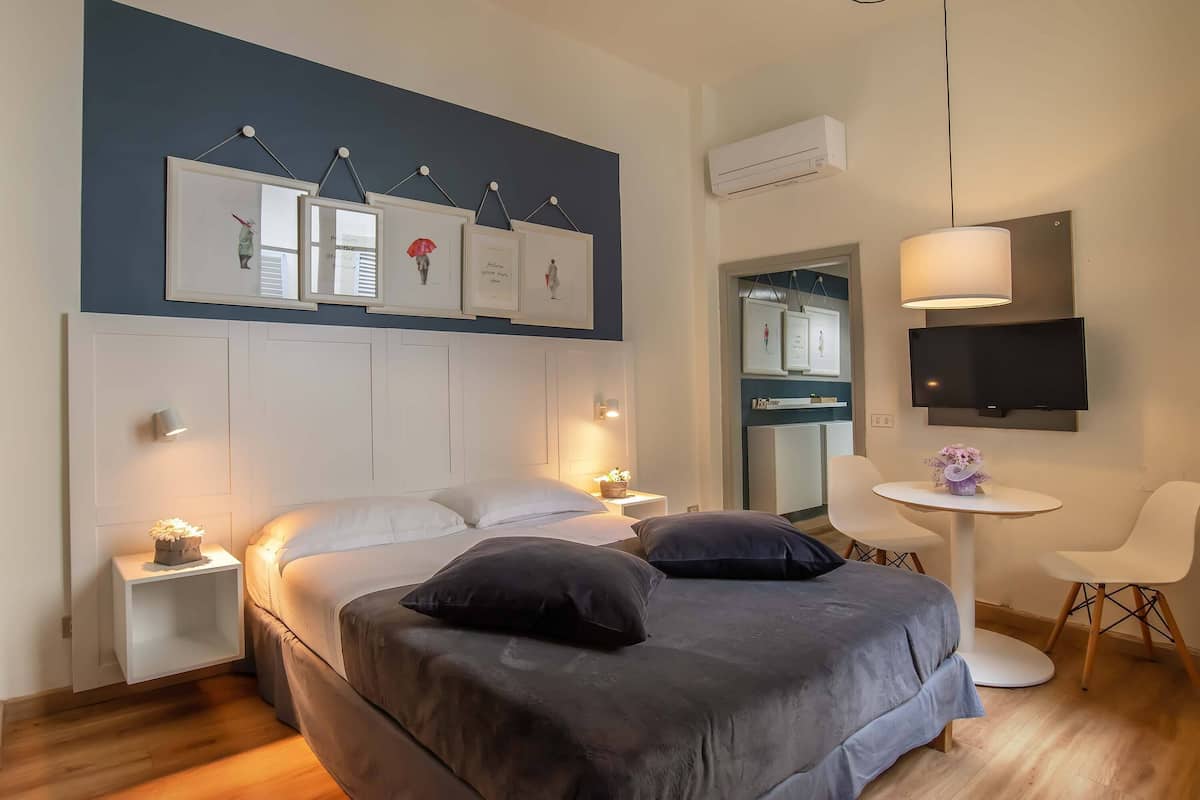 A modern hotel room with a bed, bedside tables, a TV, a small table with chairs, and artwork on the wall.