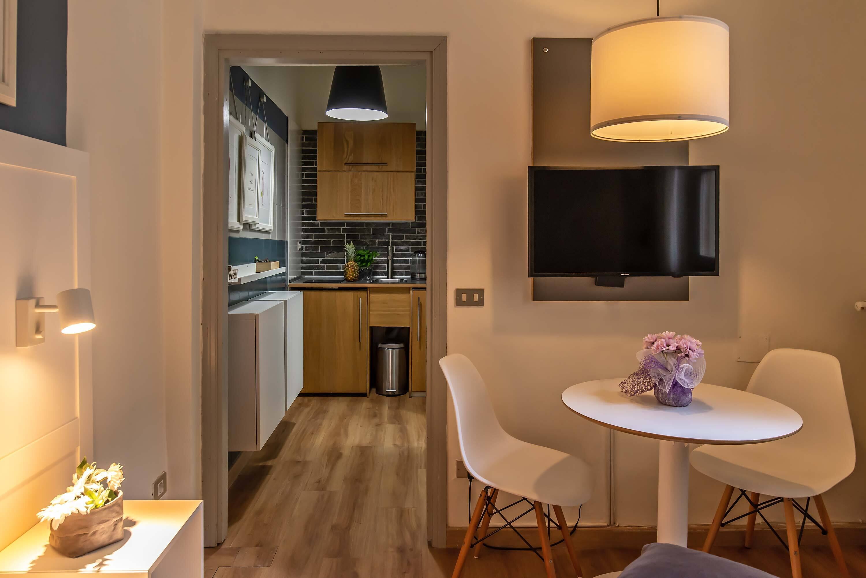 A modern kitchen with wooden cabinets, a TV mounted on the wall, and a dining area with a white table and chairs.
