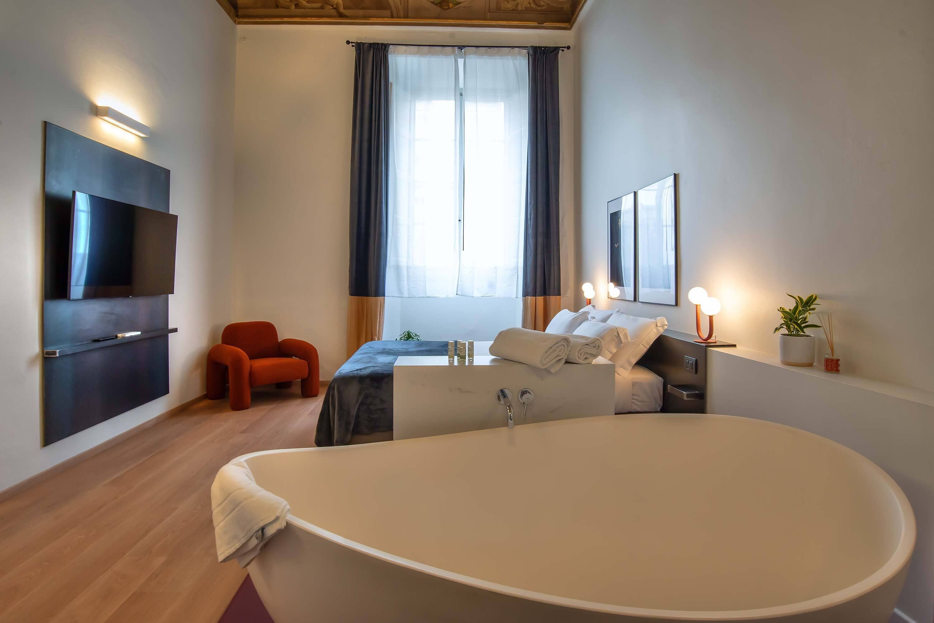 Una habitación de hotel moderna con una bañera grande, un televisor de pantalla plana montado en la pared, un sillón cómodo y una cama con ropa de cama impecable.