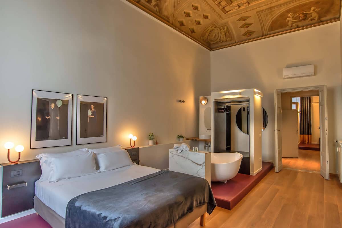 A hotel room with a bed, bedside tables, a vanity with a mirror, a large white bathtub, and a ceiling with decorative elements.