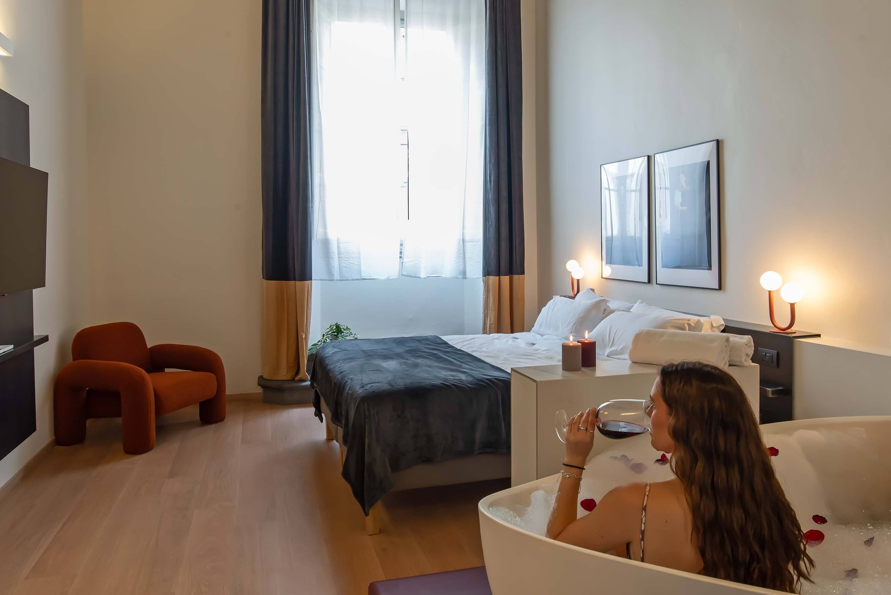 A modern hotel room with a bed, a sitting area with a chair, a bathtub with rose petals, and a large window with curtains.