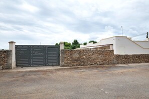 Una entrada con reja y un muro de piedra, un edificio blanco con techo plano, y cielo despejado.