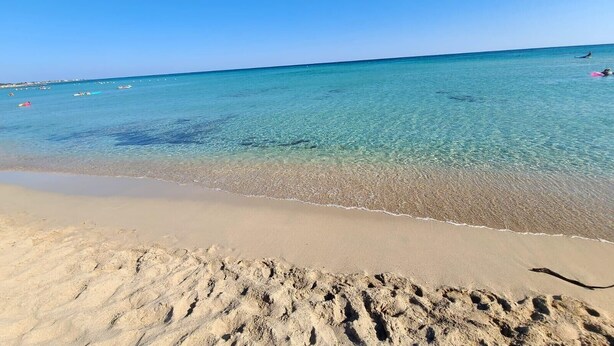 Una playa con agua cristalina, una orilla de arena y barcos a lo lejos.