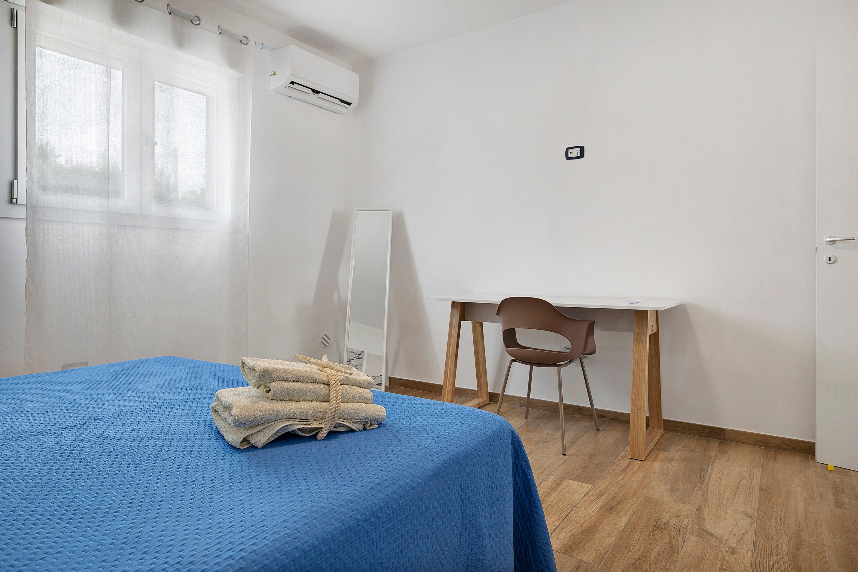 A room with a bed, a desk, and a chair. There is a window with curtains, an air conditioning unit, and a power outlet on the wall.