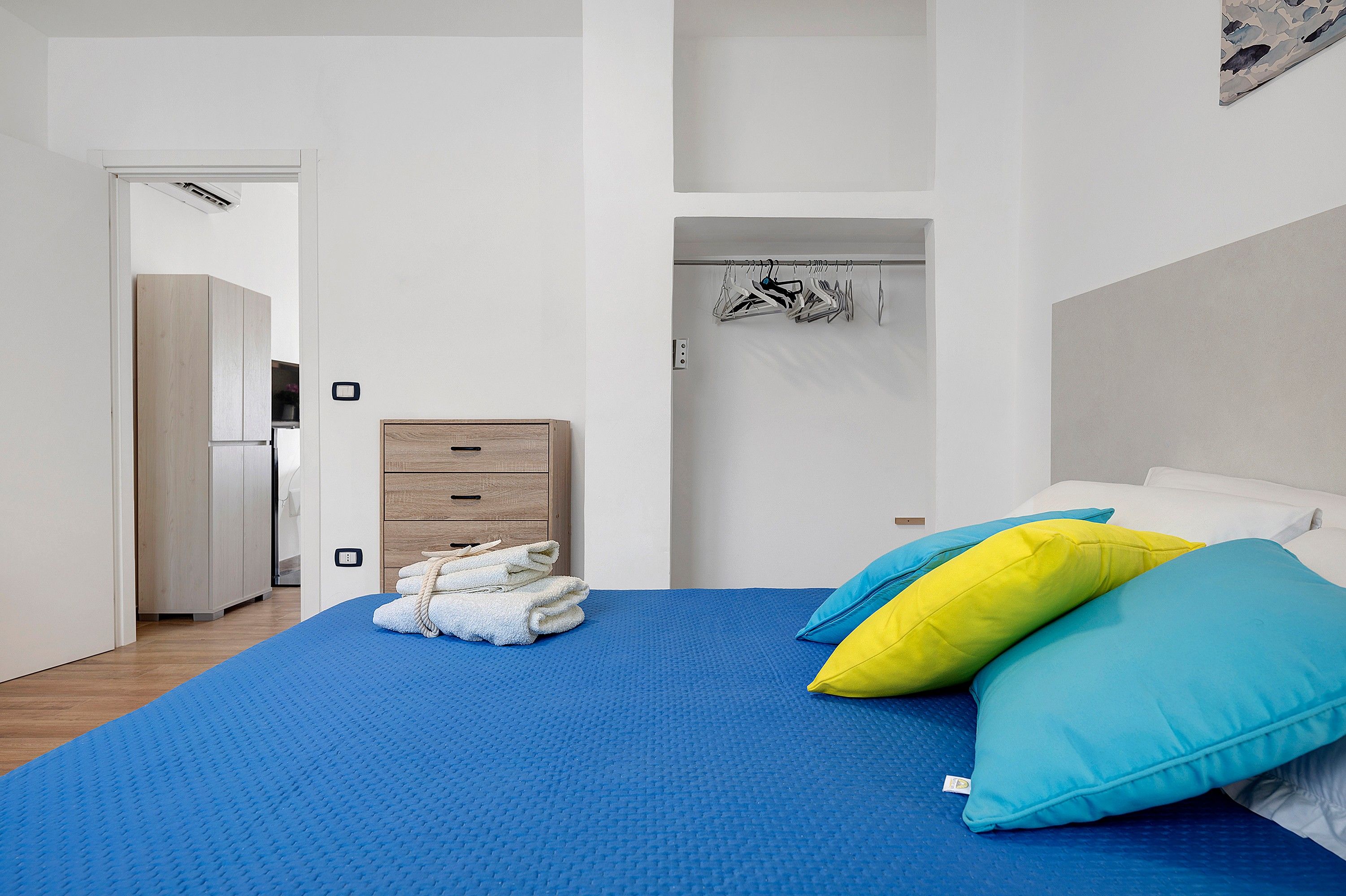 A bedroom with a bed, a wooden dresser, a hanging clothes rack, and a door leading to another room.