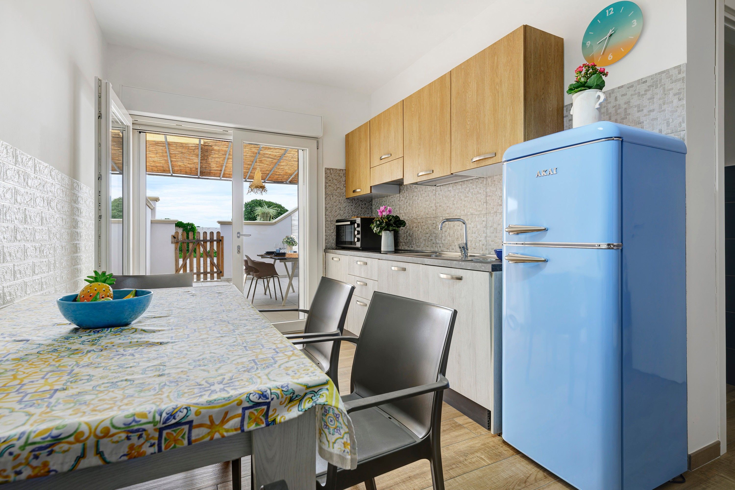 A compact kitchen with wooden cabinets, a blue refrigerator, and a dining area with a patterned tablecloth.