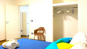 Una camera d'hotel con un letto, una sedia, un appendiabiti e una porta che dà su un'altra stanza.