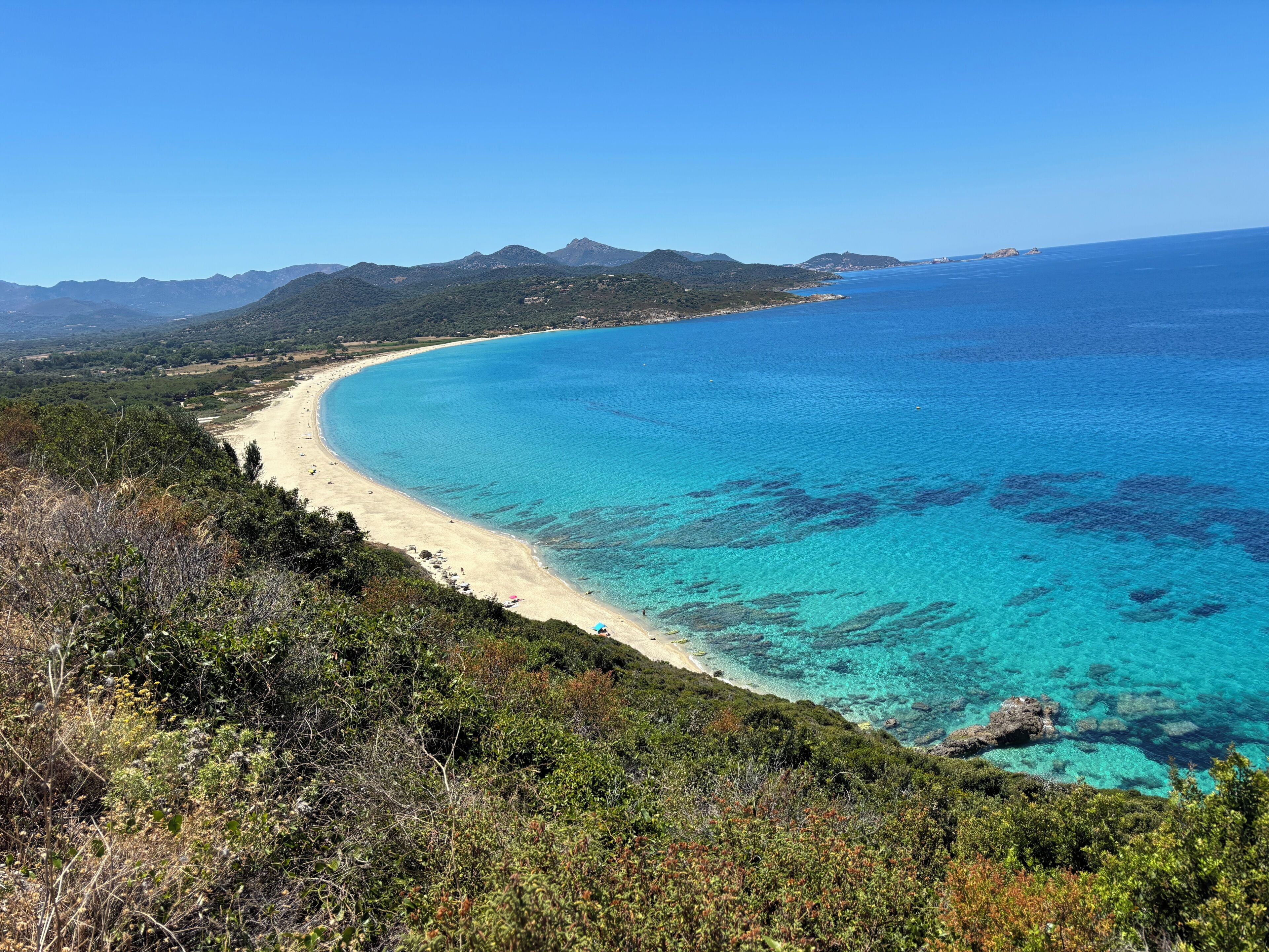 Eine Küstenlandschaft mit Strand, klarem blauen Wasser und umliegenden Hügeln.