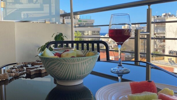 Een tafel met een schaal fruit, een glas rode wijn en een bord met watermeloen op een balkon met uitzicht over de stad.