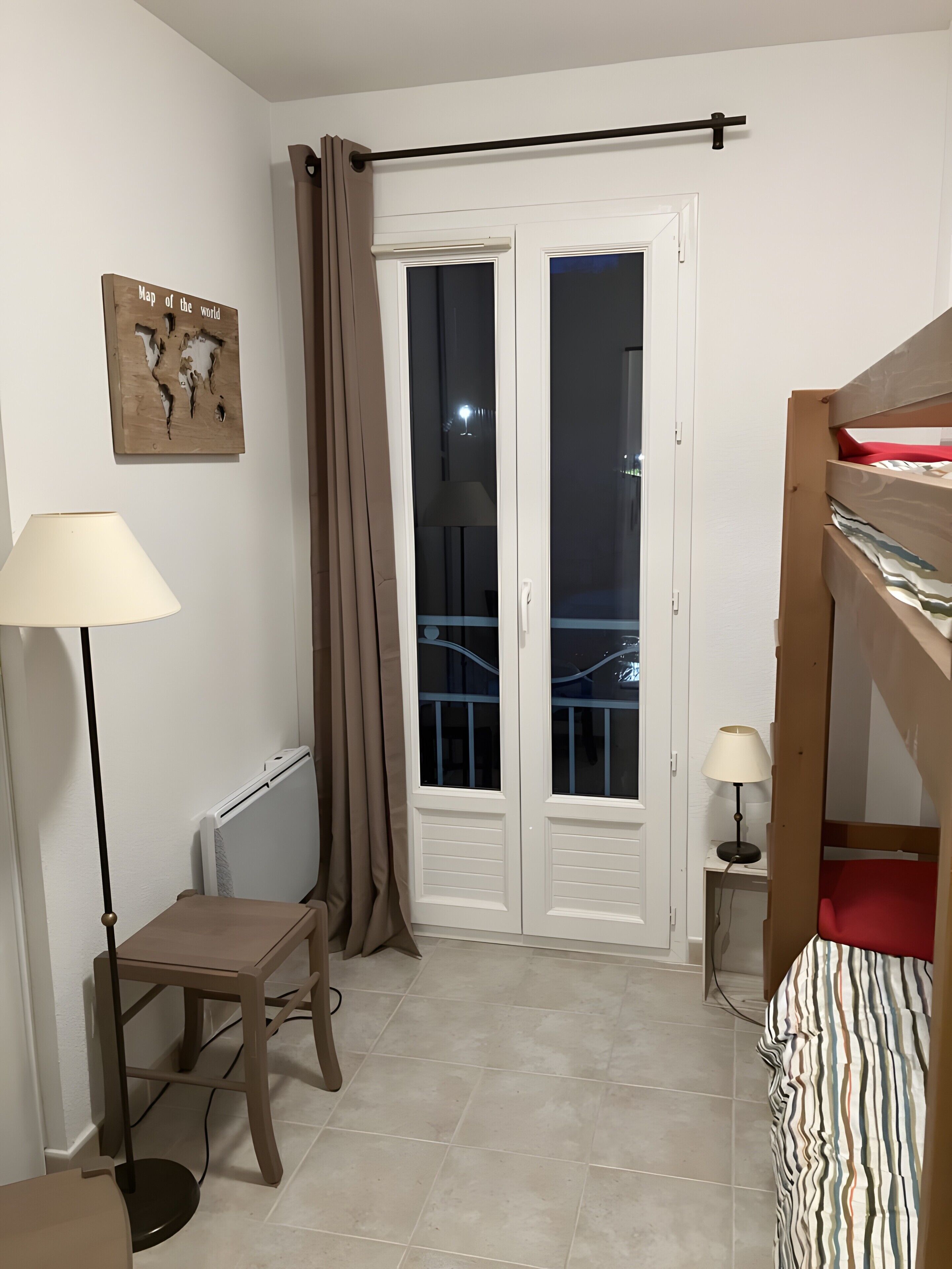 Una habitación pequeña con cama, una silla, una mesa, radiador, lámpara, un mapa en la pared y un balcón con cortina.