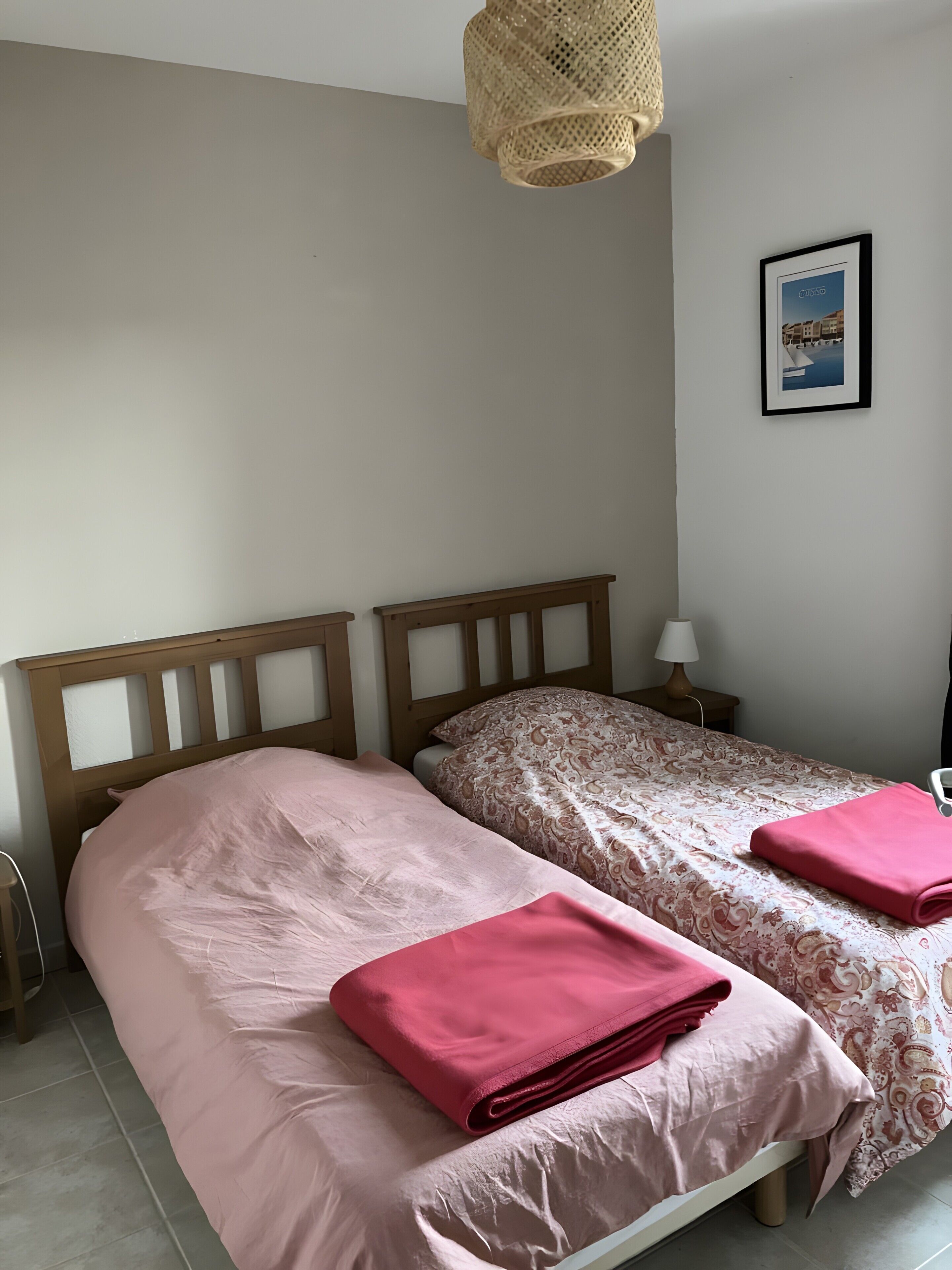 Un dormitorio con dos camas, una lámpara de noche, un cuadro enmarcado en la pared y una luminaria tejida.