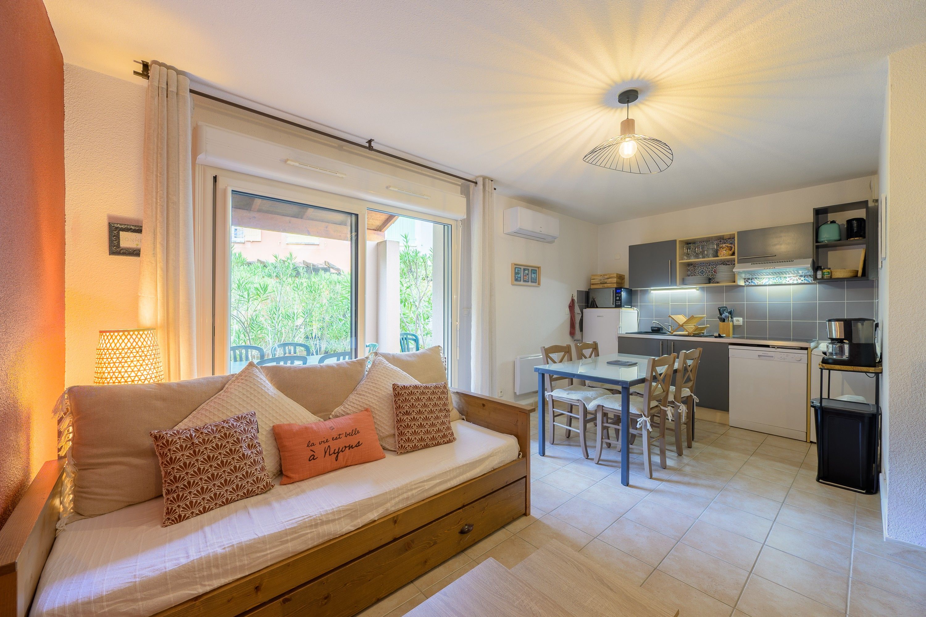 Un salón acogedor con sofá, una mesa de comedor y sillas, una cocina con electrodomésticos modernos, y vistas al exterior con vegetación.