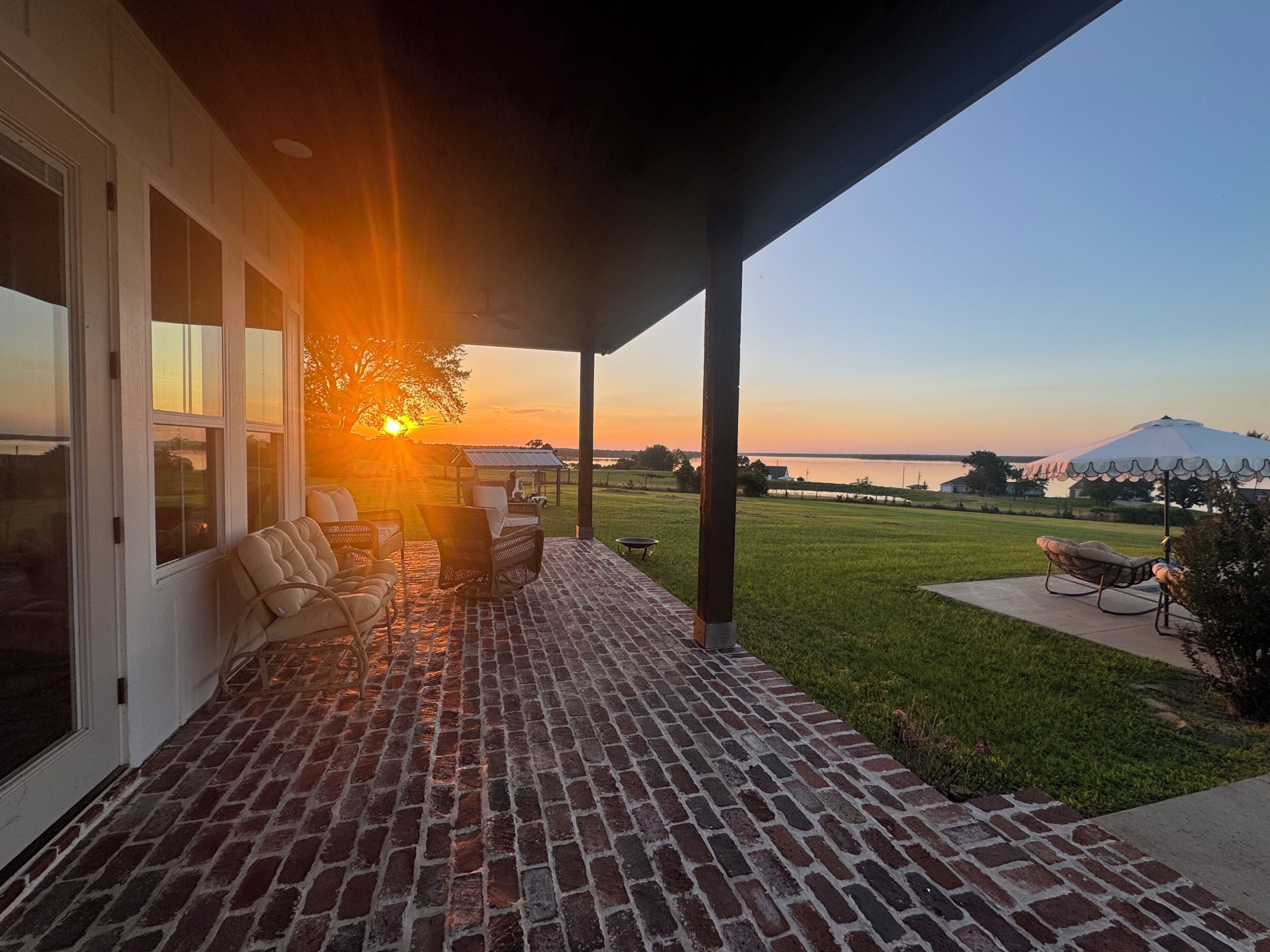 A covered patio with brick flooring, outdoor furniture, and a view of a body of water at sunset.