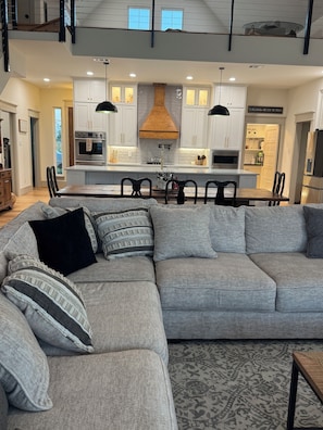 A modern kitchen with white cabinets, a central island, and a dining area with a grey sectional sofa.